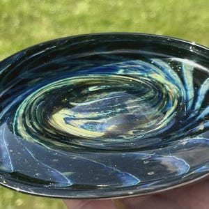 May include: A hand-blown glass bowl with a swirling galaxy design. The bowl features a dark blue exterior with interior swirls of blue, green, and yellow. The bowl is approximately 6 inches in diameter.