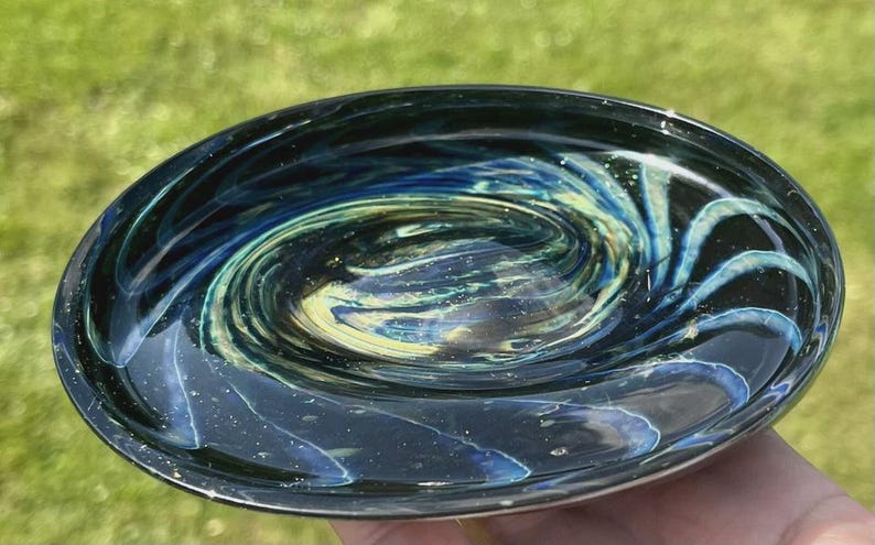 May include: A hand-blown glass bowl with a swirling galaxy design. The bowl features a dark blue exterior with interior swirls of blue, green, and yellow. The bowl is approximately 6 inches in diameter.