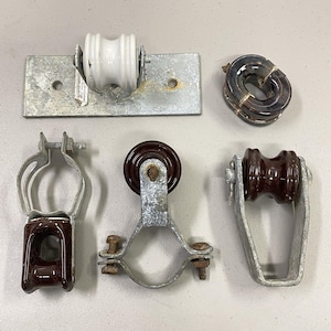 May include: Collection of vintage electrical insulators and hardware. Includes a white ceramic pulley on a metal plate, a brown ceramic ring insulator, and several brown ceramic insulators with metal brackets and clamps.