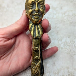 May include: A brass nutcracker shaped like a jester with a detailed face, a ruffled collar, and a long handle featuring two circular buttons and a leaf-like design at the base. The metal has a polished gold-toned finish with dark patina accents.