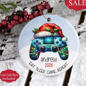 May include: A round, blue and white Christmas ornament featuring a video game controller design. The controller is decorated with Christmas lights and a Santa hat. The text "Andrew 2026" and "EAT. SLEEP. GAME. REPEAT." are also on the ornament.