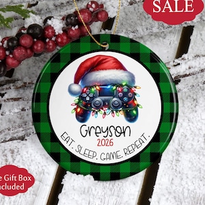 May include: A round ceramic Christmas ornament featuring a green and black buffalo plaid border. The center displays a video game controller wearing a Santa hat and wrapped in colorful string lights, with the text Greyson 2026 Eat. Sleep. Game. Repeat.