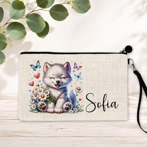 May include: A beige canvas zippered pouch with a black wrist strap. The front features a graphic of a smiling puppy holding a bouquet of white daisies, surrounded by blue butterflies and red hearts. The name Sofia is printed in black script.