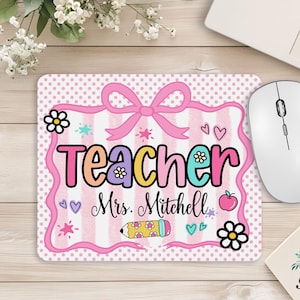 May include: A rectangular mouse pad with a pink and white striped background and polka dot border. It features a pink bow, a pencil, an apple, and flowers, with the text "Teacher" and "Mrs. Mitchell" printed in a colorful, playful font.