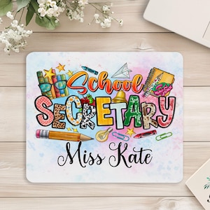 May include: A rectangular mousepad with the words "School Secretary" in colorful letters, along with the name "Miss Kate." The design includes school-themed illustrations like books, pencils, and a bell, set against a watercolor background.