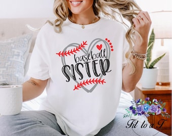 May include: A white short-sleeved t-shirt featuring a graphic design of a grey heart outlined with red baseball stitching. The text "baseball SISTER" is printed in black script and bold lettering across the center of the heart, accompanied by a small red heart icon.