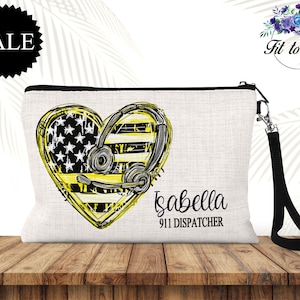 May include: A white canvas zippered pouch with a black strap. The pouch features a yellow and black heart with an American flag design and a headset. The text "Isabella 911 Dispatcher" is printed below the heart.