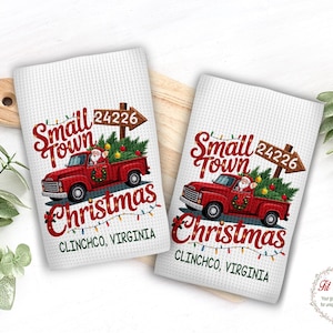May include: Two white waffle-textured kitchen towels featuring a red pickup truck carrying a Christmas tree. The design includes the text Small Town Christmas, the number 24226, and Clinchco, Virginia, accented with colorful holiday string lights.