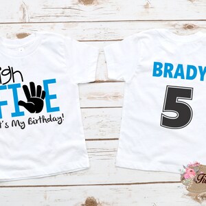 May include: Two white t-shirts with blue and black text. The front of one shirt says "high FIVE" with a black handprint and "It's My Birthday!" The back of the other shirt says "BRADY 5" in blue and black text.
