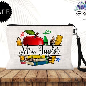 May include: A white canvas cosmetic bag with a black zipper and wrist strap. The bag features a red apple, pencils, crayons, and books, with the text "Mrs. Taylor" and a "SALE" tag.