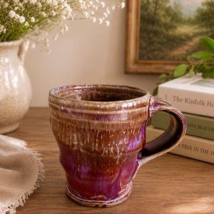 May include: A ceramic mug with a glossy finish featuring a blend of deep purple and earthy brown tones. The mug has a cylindrical body with a slightly flared rim and a sturdy, curved handle. The glaze shows a dripped, textured effect throughout.