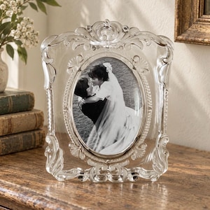 May include: A clear, ornate glass picture frame with a rectangular outer edge and a central oval opening. The frame features intricate scrollwork and relief detailing along the borders. It holds a black and white photograph of two people embracing.