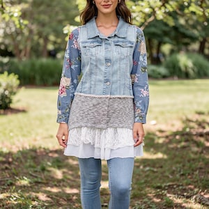 May include: A light-wash denim vest with a button-up front, layered over a floral-print blouse and a gray and white lace-trimmed tunic. The outfit is completed with light-wash denim jeans, creating a layered, casual look.