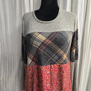 May include: A long-sleeved top with a color-block design. The top features a gray knit upper, a plaid middle section in gray, tan, and pink, and a red and pink patterned lower section. The top has a round neckline and button details.