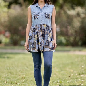 May include: A sleeveless denim top with floral patchwork detailing, paired with blue jeans. The top features a collared neckline and button-down front. The patchwork incorporates shades of blue, yellow, and brown. The model is wearing blue jeans and beige slip-on shoes.