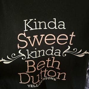 May include: A black t-shirt featuring white and light pink text that reads Kinda Sweet Kinda Beth Dutton Yellowstone, with decorative flourishes surrounding the word kinda. The shirt has a black and white plaid pattern visible at the neckline.