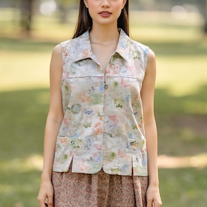 May include: Sleeveless vest with a collared neckline and a front zipper. The vest features a beige background with a colorful floral and butterfly pattern. It has two front pockets with button closures. The model is also wearing a brown patterned skirt.