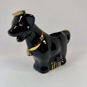 May include: Black ceramic dog figurine with gold accents. The dog has a collar and a hat, and a small opening on its back. The figurine is a decorative item.