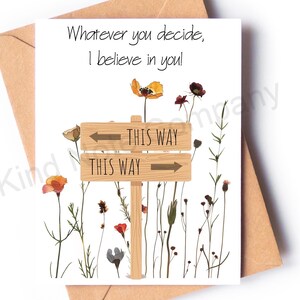 May include: A white card with a message that reads "Whatever you decide, I believe in you!"  The card features a wooden sign with two arrows pointing in opposite directions. The sign reads "This Way" on both sides. The card is decorated with a floral design.