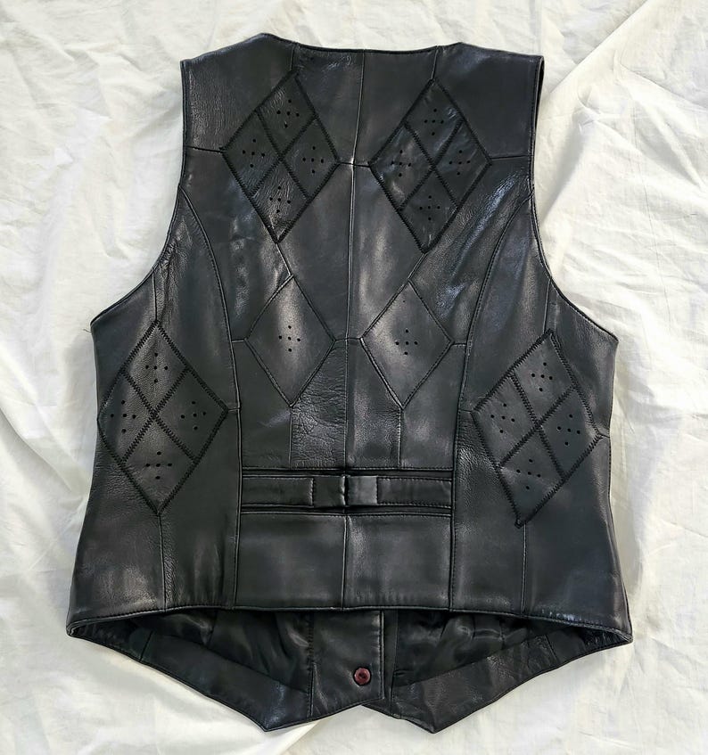 Puede incluir: Chaleco de cuero negro con detalles en forma de diamante y perforaciones. El chaleco tiene cierre de bot&oacute;n y un corte entallado. La parte trasera del chaleco es visible, mostrando las intrincadas costuras y el dise&ntilde;o. Una prenda de vestir cl&aacute;sica.