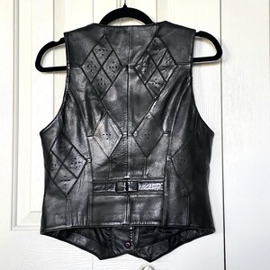 Puede incluir: Chaleco de cuero negro con recortes en forma de diamante y dise&ntilde;o de patchwork. El chaleco tiene cierre de bot&oacute;n y est&aacute; colgado de una percha negra. Una prenda de vestir cl&aacute;sica.
