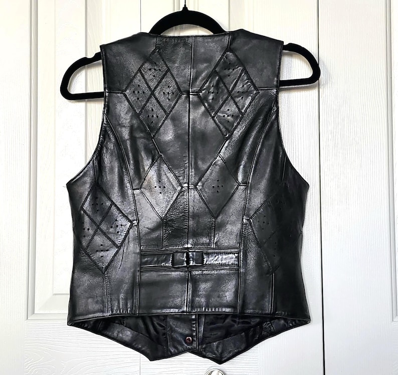 Puede incluir: Chaleco de cuero negro con recortes en forma de diamante y dise&ntilde;o de patchwork. El chaleco tiene cierre de bot&oacute;n y est&aacute; colgado de una percha negra. Una prenda de vestir cl&aacute;sica.