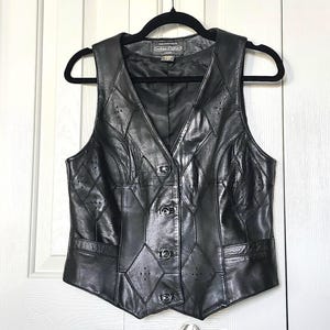 Puede incluir: Chaleco de cuero negro con cuello en V y cierre de botones. El chaleco presenta un dise&ntilde;o de patchwork con recortes en forma de diamante y costuras decorativas. El chaleco sin mangas cuelga de una percha negra.