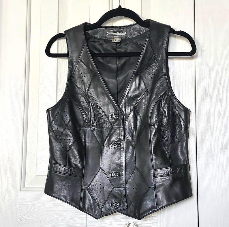 Puede incluir: Chaleco de cuero negro con cuello en V y cierre de botones. El chaleco presenta un dise&ntilde;o de patchwork con recortes en forma de diamante y costuras decorativas. El chaleco sin mangas cuelga de una percha negra.