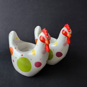 May include: Two ceramic chicken-shaped candle holders. The white chickens are decorated with colorful polka dots in orange, green, pink, and yellow. They have red combs and beaks, and are set against a dark background.