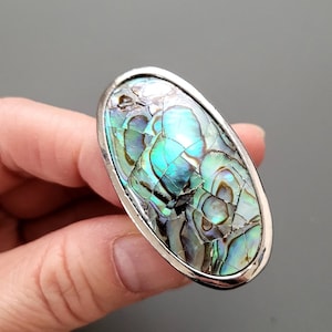 May include: Oval shaped ring with a silver setting and a large iridescent abalone shell inlay. The shell has a mosaic pattern of green, blue, and brown.