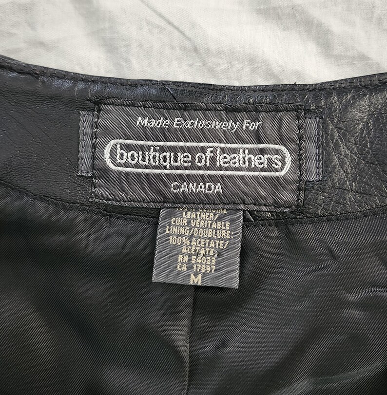 Puede incluir: Primer plano de una etiqueta de prenda de cuero negro. La etiqueta dice "Made Exclusively For boutique of leathers CANADA". La etiqueta tambi&eacute;n enumera los materiales como cuero y 100% acetato. La talla es M.