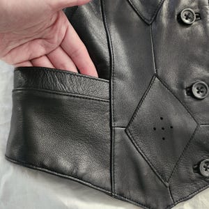 Puede incluir: Chaleco de cuero negro con dise&ntilde;o geom&eacute;trico y tres botones decorativos. El chaleco tiene un bolsillo en el lado izquierdo. El cuero parece suave y flexible.