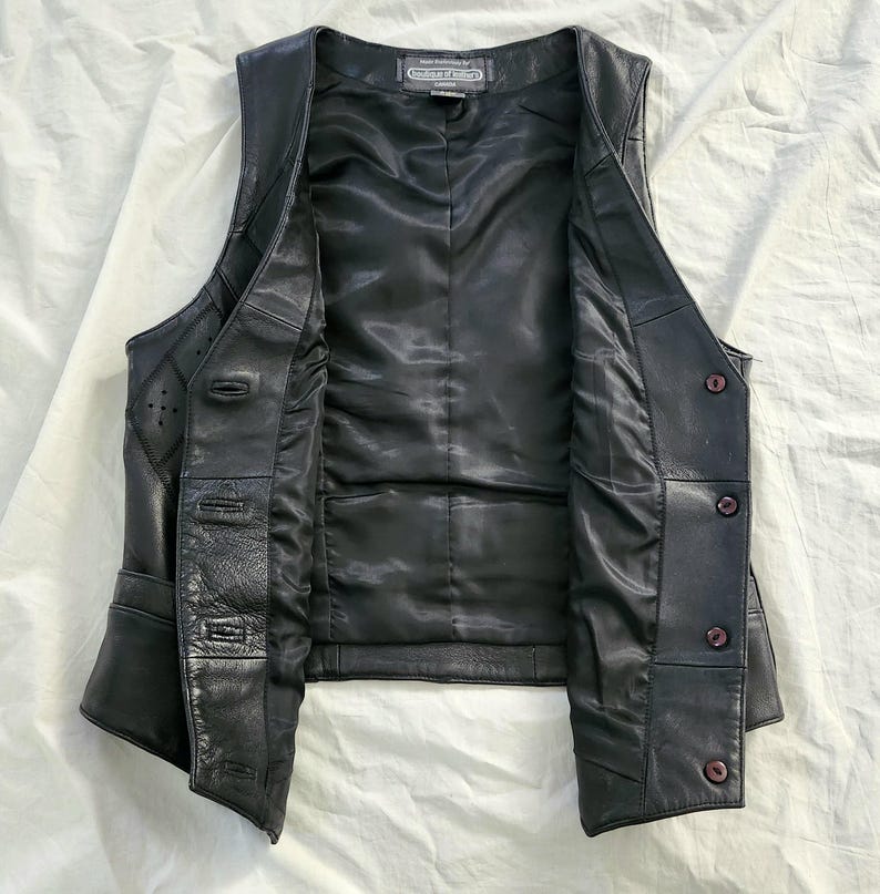 Puede incluir: Chaleco de cuero negro con cierre de botones y forro de sat&eacute;n. El chaleco tiene un dise&ntilde;o cl&aacute;sico con un corte entallado, costuras decorativas y detalles sutiles. El chaleco est&aacute; abierto, mostrando el forro interior y los cierres de bot&oacute;n.