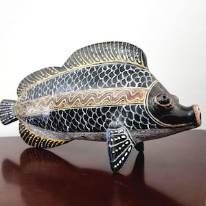 May include: A hand-carved, decorative fish sculpture. The fish is painted black with white scale detailing and gold and red accents. The sculpture has a detailed fin and tail design, and is resting on a dark brown surface.