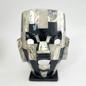 May include: A decorative mask-shaped vase with a geometric design. The vase is crafted from black, white, and iridescent materials, creating a striking visual contrast. The mask features a square base and a stylized face with open eye and mouth shapes.