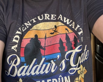 Op de afbeelding: Een zwart T-shirt met een retro-grafisch ontwerp met een zonsondergang, een personage met een zwaard en de tekst "Adventure Await Baldur's Gate Faerun Sword Coast".