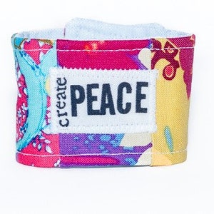 Op de afbeelding: Kleurrijke stoffen koffiemouw met een patchwork ontwerp in roze, blauw en geel. De mouw heeft de tekst "create PEACE" op een wit label. Ontworpen voor een standaard koffiekopje.