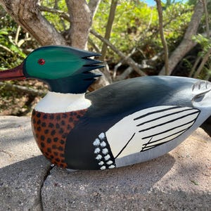 May include: Hand-painted wooden duck figurine. The duck features a green head, red beak, and a body with black, white, and brown details. The wings are painted with white and black stripes. The duck is resting on a stone surface.