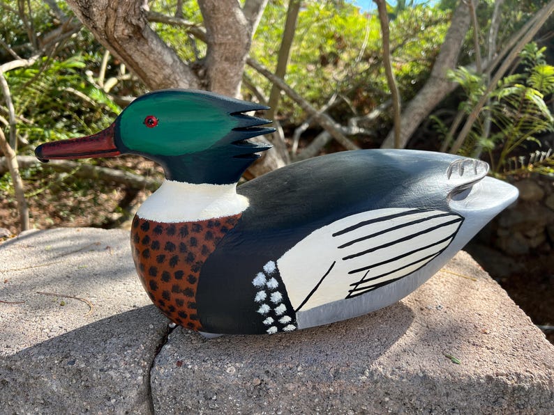May include: Hand-painted wooden duck figurine. The duck features a green head, red beak, and a body with black, white, and brown details. The wings are painted with white and black stripes. The duck is resting on a stone surface.
