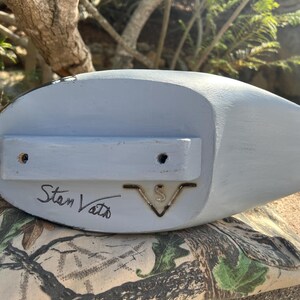 May include: A light blue wooden duck decoy with a flat bottom and a rounded top. The decoy has a rectangular wooden piece attached to the bottom with two holes. The name "Stan Vath" is handwritten on the bottom.