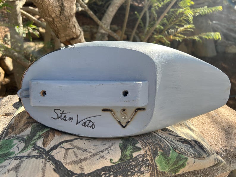 May include: A light blue wooden duck decoy with a flat bottom and a rounded top. The decoy has a rectangular wooden piece attached to the bottom with two holes. The name "Stan Vath" is handwritten on the bottom.
