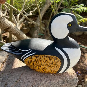 May include: A hand-painted wooden duck sculpture. The duck has a white head, black and white body, and a yellow and orange patterned chest. The sculpture is on a stone, with a natural outdoor background.