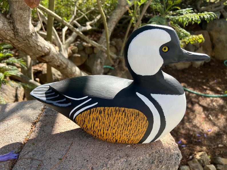 May include: A hand-painted wooden duck sculpture. The duck has a white head, black and white body, and a yellow and orange patterned chest. The sculpture is on a stone, with a natural outdoor background.