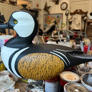 May include: A hand-painted wooden duck decoy. The duck has a black body, white head markings, and a yellow and brown textured breast. It features a black beak and yellow eye. Decoys like this are used for hunting or as decorative items.
