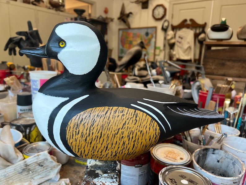 May include: A hand-painted wooden duck decoy. The duck has a black body, white head markings, and a yellow and brown textured breast. It features a black beak and yellow eye. Decoys like this are used for hunting or as decorative items.