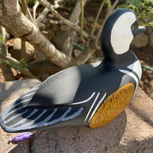 May include: A hand-painted wooden duck figurine. The duck has a black body, white head with a black beak, and a yellow and brown wing. The duck is on a stone surface, with a natural background.