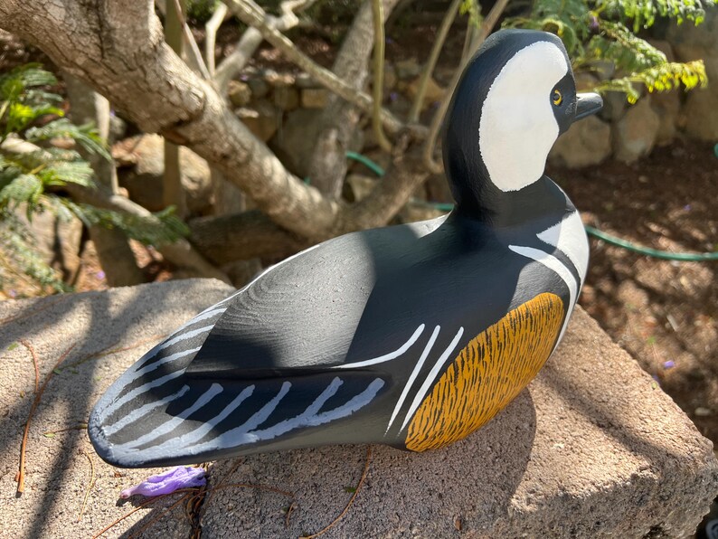 May include: A hand-painted wooden duck figurine. The duck has a black body, white head with a black beak, and a yellow and brown wing. The duck is on a stone surface, with a natural background.