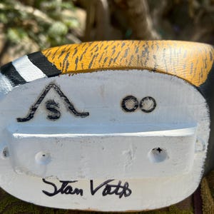 May include: Close-up of a hand-painted wooden duck decoy. The decoy features a white base with black and yellow accents. The text "Stan Vath" is hand-written in black ink on the bottom of the decoy.