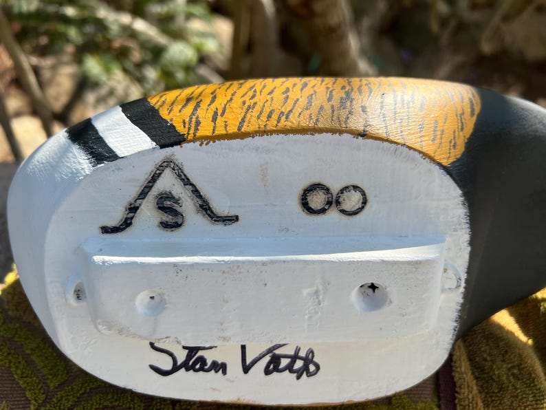 May include: Close-up of a hand-painted wooden duck decoy. The decoy features a white base with black and yellow accents. The text "Stan Vath" is hand-written in black ink on the bottom of the decoy.