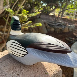 May include: A hand-painted wooden duck figurine. The duck has a green head, black and white neck, and a gray and brown body. The duck is resting on a stone surface.